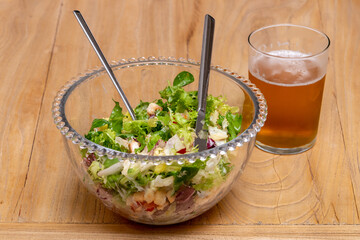 a glass bowl with a Caesar salad with lettuce, tomato, chicken breast, sauce and crusty bread, with a glass of cold beer with foam, a fork and a stabbed knife, on a wood table