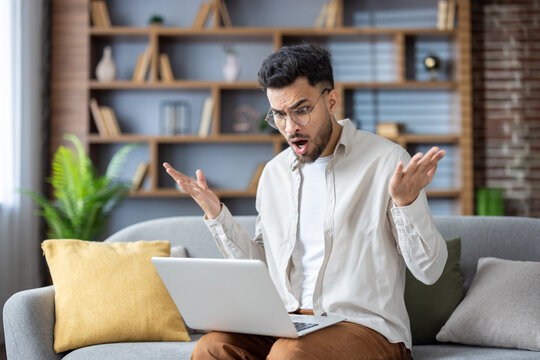 Shocked and worried young muslim man man sitting on sofa at home and dissapointed spreading his hands looking at laptop screen