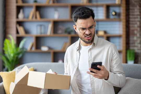 Angry young Indian man sitting at home holding mistakenly received package and looking worriedly at mobile phone screen