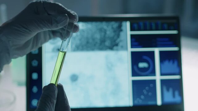 Close-up of hands of unrecognizable scientist in gloves studying test tube with green liquid chemical solution against light from computer screen for residue, while conducting experiment in laboratory