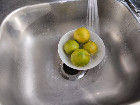 Un primer plano de varias naranjas verdes y amarillas en un plato de color blanco cay&eacute;ndole agua dentro de un fregadero en la cocina.
