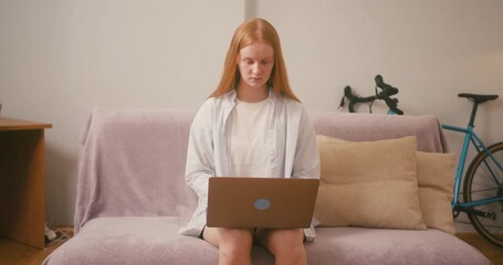 Red-haired woman types on laptop comfortably sitting on sofa - Powered by Adobe