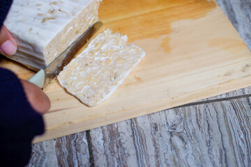 Close up Female Hand Slicing Cut Tempeh or Tempe on Wooden Chopping Board Using Knife. Tempe is Fermented Soy Product Originally from Indonesia.