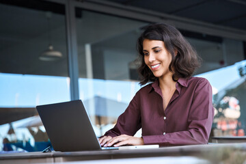 Smiling middle eastern indian woman working remotely online on business project. Young hispanic or muslim freelance female student worker using laptop for studying sitting outdoors at office building