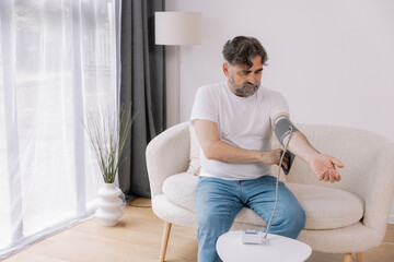 Handsome grey-haired senior man in casual outfit sitting on couch at home, checking blood pressure...