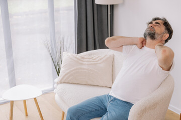A senior man is sitting indoors, visibly suffering from neck pain. He is holding his neck with a pained expression, indicating discomfort, copy space for your advertising