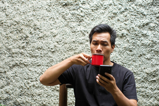 A Southeast Asian man wearing black t shirt sitting on bamboo chair in the terrace, drinking black coffee on red mug and holding smartphone, relax time. - Powered by Adobe
