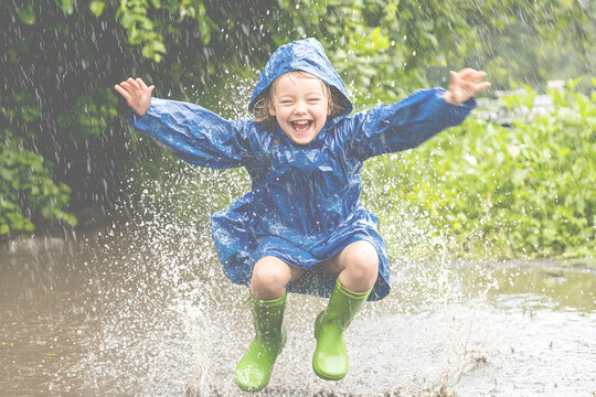 Happy child in a blue raincoat and green boots jumping in a puddle on a rainy day