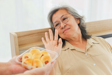 Elderly asian woman raising her hand refuse eat sweet sausage  bakery snacks containing flour and sugar that are harmful to her health while she was sick in bed : Concept eating nutritious food.