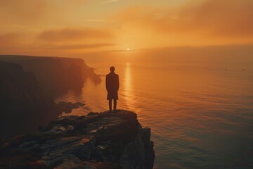 Lonely man silhouette stands on rocky cliff, watching vibrant sunset over ocean, enveloped in warm glowing hues, capturing moment of quiet contemplation and peace