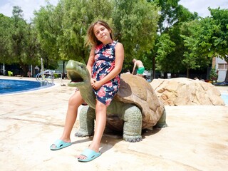 Young girl sitting on a large turtle statue by the pool at a water park on a sunny day
