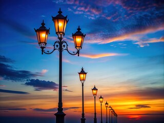 Obraz premium Dark silhouettes of slender lamp posts stretch towards the horizon, contrasting elegantly against a serene, vibrant blue twilight sky with a hint of warm sunset tones.