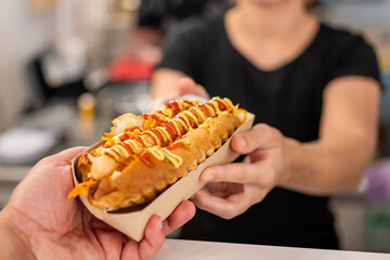 Close-up of a hand holding a hot dog with mustard and ketchup, with an out-of-focus person in the...