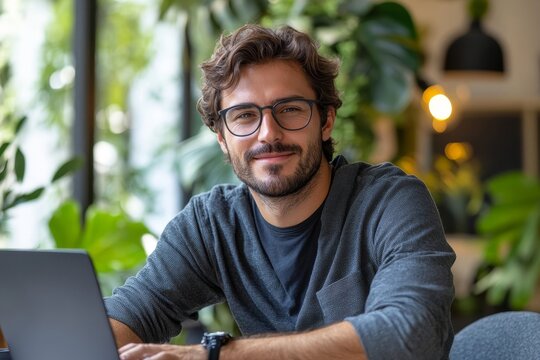 Portrait of a young man in glasses sitting at a desk in the office and working on a laptop, looking and smiling at the camera, Generative AI - Powered by Adobe