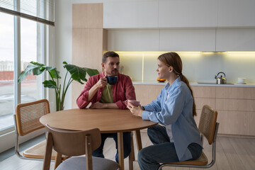 Thoughtful husband with interested woman talking together, sitting on kitchen, drinking morning...