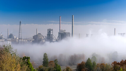 Industrial factory plant with tall smokestacks emitting thick smoke, enveloped in heavy fog. The image highlights the impact of industrial pollution on the environment