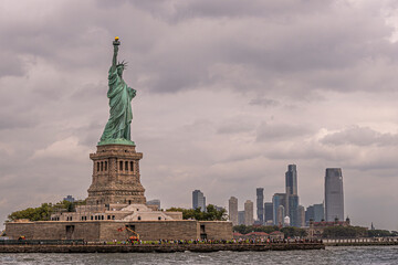 Fototapeta premium New York, NY, USA - August 4, 2023: Statue of Liberty from the bay plus New Jersey City skyline under gray cloudscape