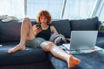Portrait of red-haired young woman sitting on sofa using laptop, grey cat sleeping nearby