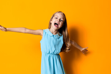 Joyful little girl in a blue dress with outstretched arms against a vibrant orange background,...
