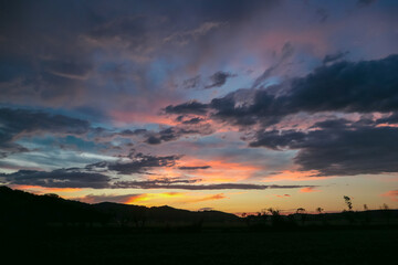 Dramatic sky in Jennersdorf, Burgenland, Austria. Thick swirling clouds painted in hues of gray, orange, pink. Sunset of warm glow creating breathtaking spectacle. Silhouette of rolling hill landscape