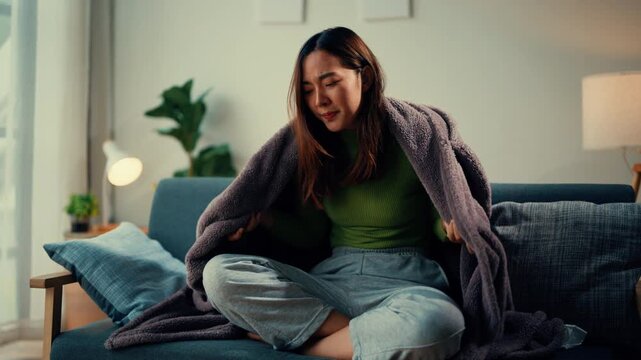 Woman feeling cold indoors wraps herself in a cozy blanket while sitting on a sofa, shivering and seeking warmth. Experience the comfort and coziness of home during the cold season
