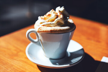 White coffee cup with whipped cream on top sits on a wooden table. Hot coffee adorned with fluffy whipped cream