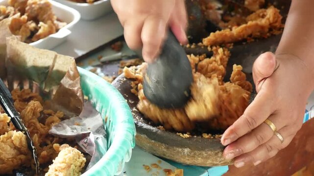 Making ayam geprek - Indonesian street food. popular fusion dish of smashed southern crispy fried chicken with topping of red chili relish. Smashing crispy fried chicken using stone mortar.