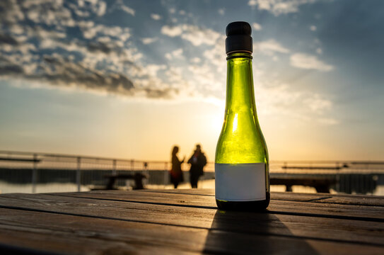 Photo of a single empty wine bottle on a wooden table, against the sunset sky, at the seaside, romantic couple silhouetted chatting in the background.