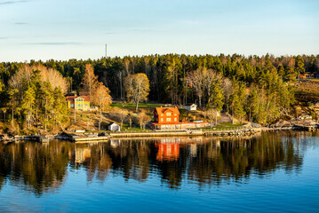 Obraz premium Small island connected with a bridge in Stockholm archipelago in Baltic sea with traditional white and red house at sunny spring evening scenery.