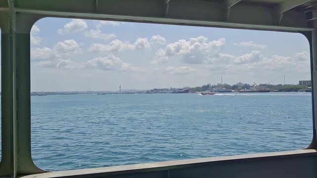Embark on a serene voyage, framed by the ferry's window, revealing a breathtaking vista of sparkling blue waters, a bustling harbor, and a picturesque cityscape under a canopy of fluf