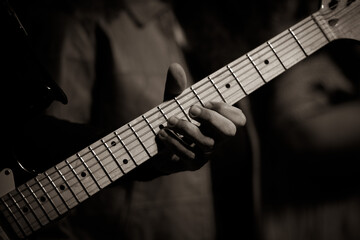 Close-up shot of a musician's hand playing an electric guitar. The black and white tones emphasize the details of the fingers on the frets and the texture of the instrument.