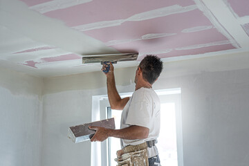 The worker make a plasterboard ceiling. He does taping plaster drywall ceiling joints. He is using special stainless steel taping knife.