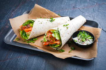 Italian style wrap piada tortilla with cured graved salmon, salad and avocado served as close-up with sour cream dressing dip on a tray