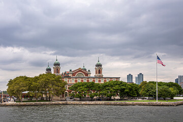 Fototapeta premium New York, NY, USA - August 4, 2023: Ellis Island National Immigration Museum shows 3 corner towers in front of 2 skylines under gray cloudscape