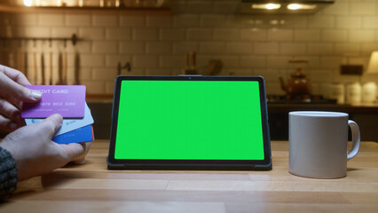 Tablet with a green screen on a kitchen counter. A person is holding multiple credit cards,...