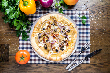 Mediterranean Pizza with black olives, meat, tomato, onion, capsicum, coriander, knife and fork served in dish isolated on napkin top view of italian fastfood on wooden table