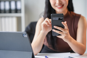Smiling asian businesswoman using smartphone in office while working on a project with a tablet on her desk