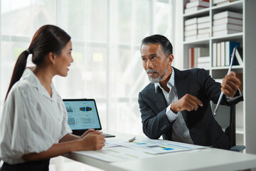 Senior manager is holding a digital tablet and explaining a business strategy to a young colleague during a meeting
