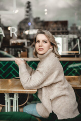 A woman is sitting at a table in a cafe, holding a cup of coffee. She is wearing a white sweater and has blonde hair. The scene is cozy and inviting