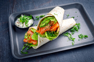 Italian style wrap piada tortilla with cured graved salmon, salad and avocado served as close-up with sour cream dressing dip on a tray
