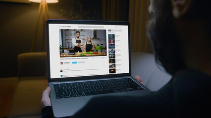 Man lying on sofa watching a food blogger couple's video on a laptop, learning how to prepare a...