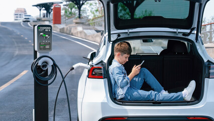 Little boy sitting on car trunk, using smartphone while recharging eco-friendly car from EV charging station. EV car road trip travel as alternative vehicle using sustainable energy concept. Perpetual