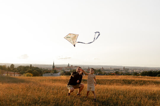 Happy father and son playing with a kite while running on meadow, sunset, in summer day. Funny family time. Happy little child launch a kite with dad.