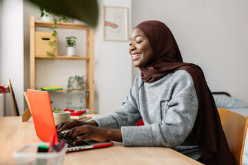 Happy black female student in muslim headscarf using laptop at home. Gen z young african woman...