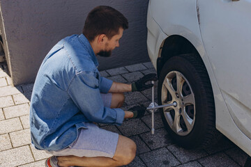 Young man in unscrewing lug nuts on car wheel in process of new tire replacement,using wrench while changing flat tire on the road.