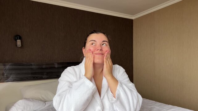 Horizontal 4K video. Young adult woman in white bath robe applies moisturizer to face in hotel room. Concept of rest, skin protection during cold season, cosmetic procedure, spa salon, relax, march 8
