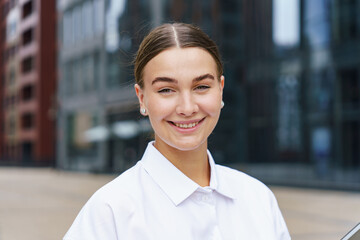 Smiling woman in a white shirt standing outdoors with a modern urban background.