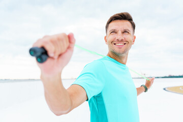 Smiling man in turquoise sportswear stretching with a resistance band outdoors on a cloudy day.