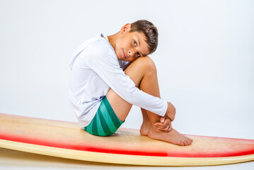 Cute barefoot boy dressed for a trip to the beach, sitting on a surfboard with his legs drawn up and his head resting on his knees