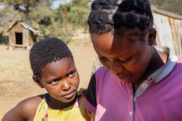 village african child girl, looking at her mother, standing in the yard in front of the shack, dog house in the background